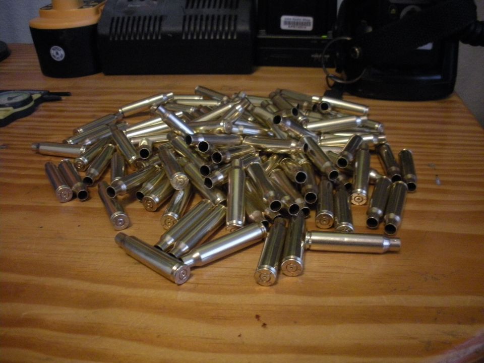 Pile of brass bullet casings on a wooden surface. Background shows electronic equipment