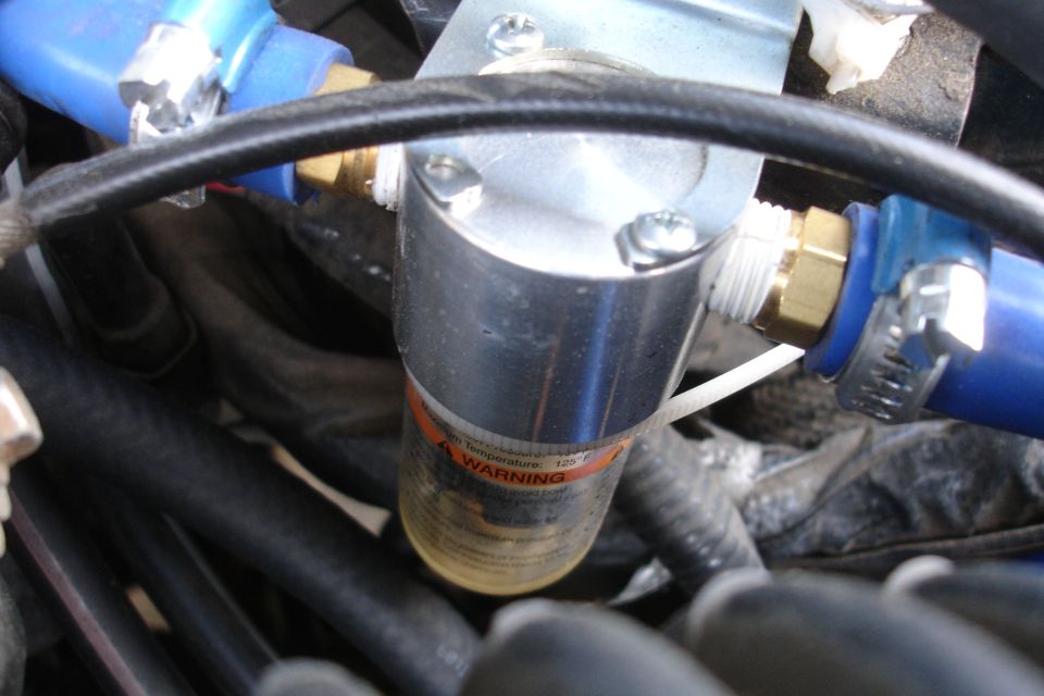 Close-up of a metal and glass engine part with blue hoses and warning label