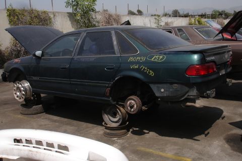 1993 Ford Taurus - A dark green 1993 Ford Taurus sits on blocks in a junkyard, missing its wheels and hood, with VIN and other markings visible on the body.