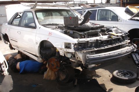 1993 Ford Taurus - Mechanic works on the front end of a disassembled 1993 Ford Taurus in a junkyard.