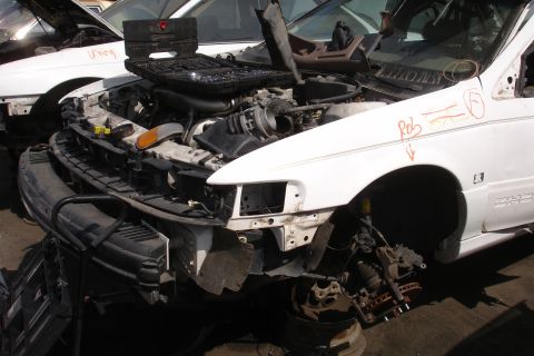 1993 Ford Taurus - A white car sits in a junkyard, its engine compartment open and front end dismantled, with another vehicle visible behind it.