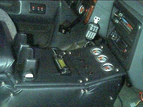 1989 Ford Bronco - Dark interior shot of a custom-built center console with a padded vinyl top, three round auxiliary gauges angled toward the driver, a CB ra…