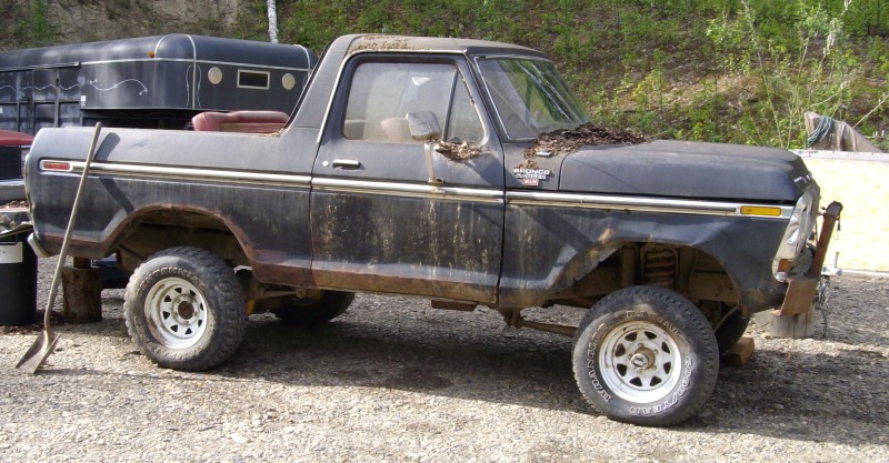 Black half-cab Ford Bronco in side view, muddy and faded, on gravel lot.