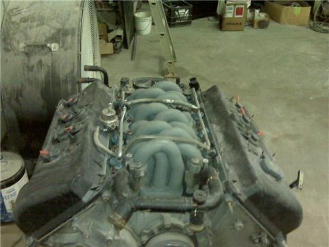 2001 Ford Crown Victoria - An engine sits on a concrete floor in a workshop, with a large fan and storage boxes nearby.