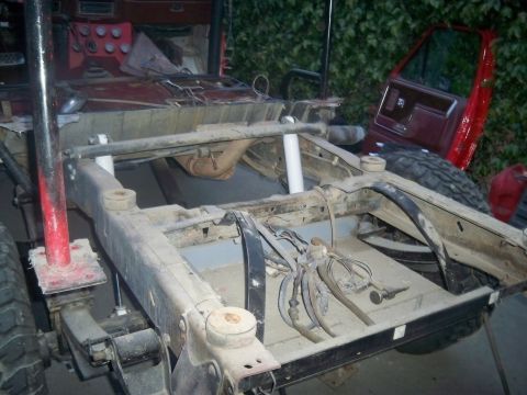 1985 Ford Bronco - The frame and undercarriage of a vehicle are shown, with various mechanical parts exposed and a detached red door nearby.