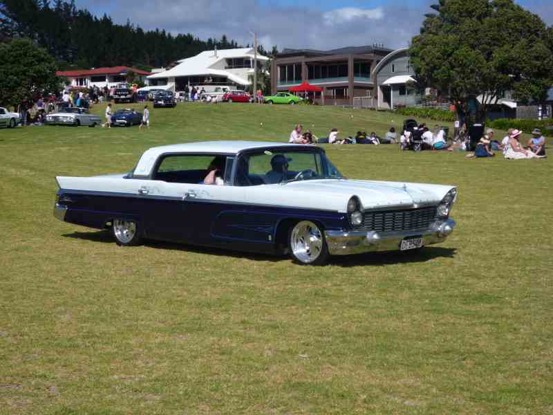 Two-tone blue and white 1958 coupe parked on grass with people in background