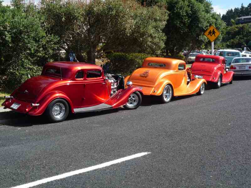 Three hot rod coupes, red, orange, and red, parked in a line on asphalt