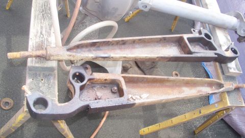 1989 Ford Bronco - Two surface-rusted radius arms lie on a shop floor after removal, showing their bushing eyes, axle-mounting brackets and threaded ends. Yel…