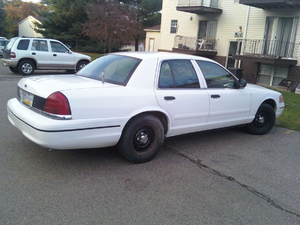 White sedan parked on asphalt, rear-left quarter view. Black steel wheels, chrome trim. Another white SUV is parked behind it