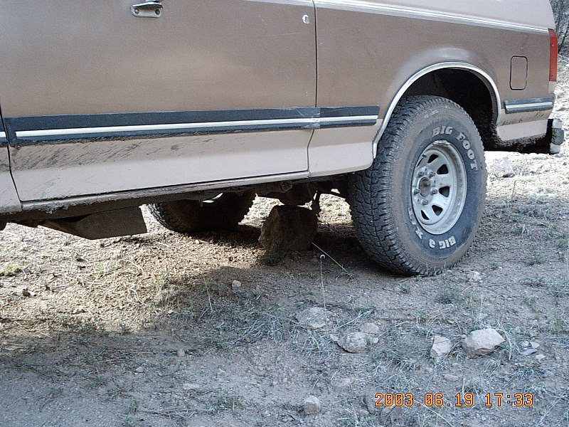 Brown Bronco side view with a rock wedged under the rear axle, Big Foot tire visible