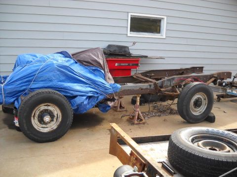 1954 Ford F-100 - A rusty truck chassis sits on jack stands, partially covered by blue tarps, with a spare tire nearby.