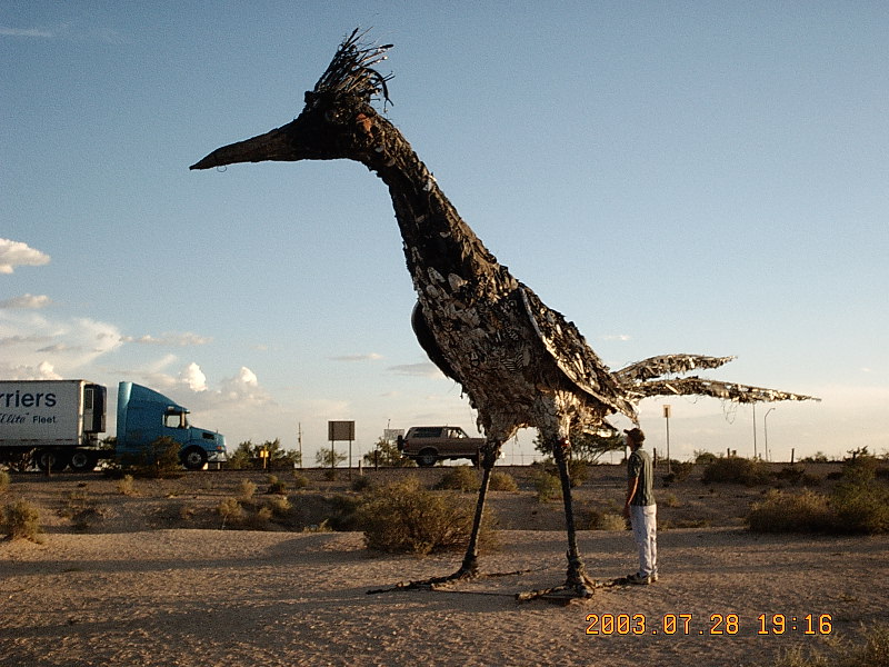 Person under huge metal roadrunner sculpture in desert; white Ford SUV and blue semi truck in background