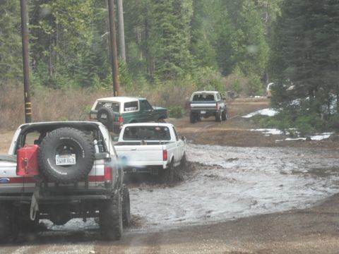 1991 Ford Bronco - Several off-road vehicles navigate a muddy path in a wooded area.
