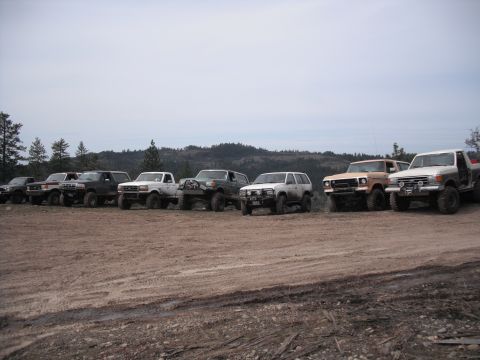 1991 Ford Bronco - Several off-road vehicles are parked in a line on a dirt lot with hills in the background.