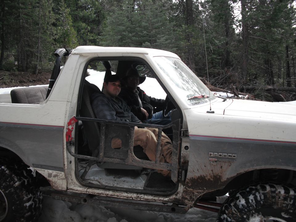 Two guys in a white 1991 Ford Bronco with the doors off, driving in snow