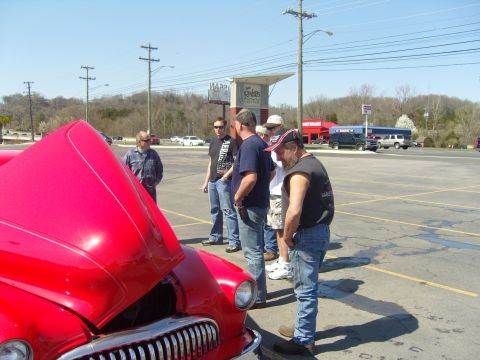 2007 Ford Mustang - A group of men inspect a classic red car with its hood raised in a parking lot.