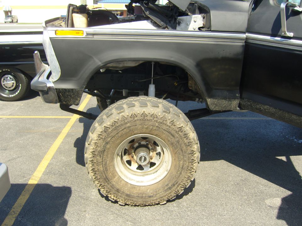 lifted pickup truck side view, showing large tire and suspension components. Another car is parked to the left