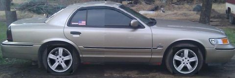 2000 Mercury Grand Marquis - A gold 2000 Mercury Grand Marquis sedan is parked outside with all four tires flat.