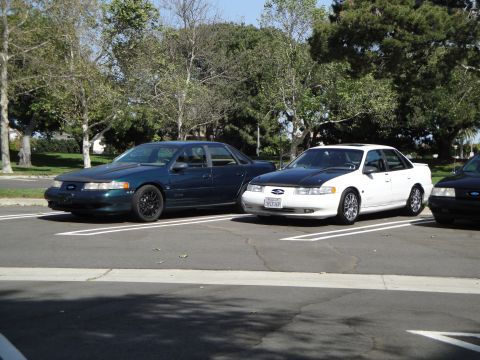 1995 Ford Taurus - Two sedans are parked in a lot, one dark green and one white with a black hood.