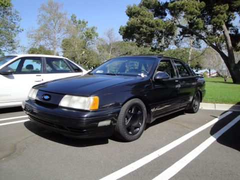1995 Ford Taurus - A black 1995 Ford Taurus is parked in a lot next to another vehicle under a clear sky.