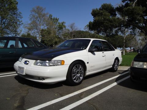 1995 Ford Taurus - A white 1995 Ford Taurus sedan is parked in a lot next to other vehicles under a clear blue sky.