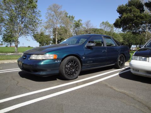 1995 Ford Taurus - A dark blue sedan with aftermarket black wheels is parked in a lot next to another vehicle.