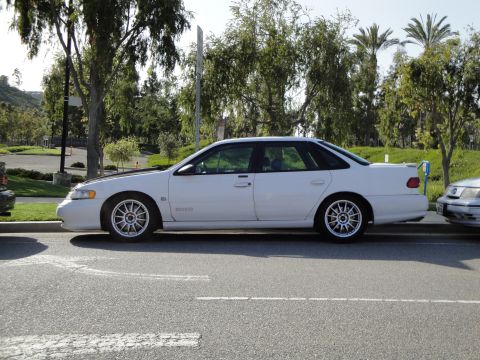 1995 Ford Taurus - Side profile of a white 1995 Ford Taurus with aftermarket wheels, parked on a street.