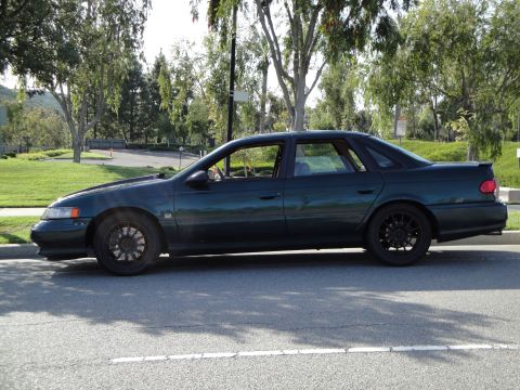 1995 Ford Taurus - A dark green 1995 Ford Taurus is parked curbside on a sunny day with trees in the background.