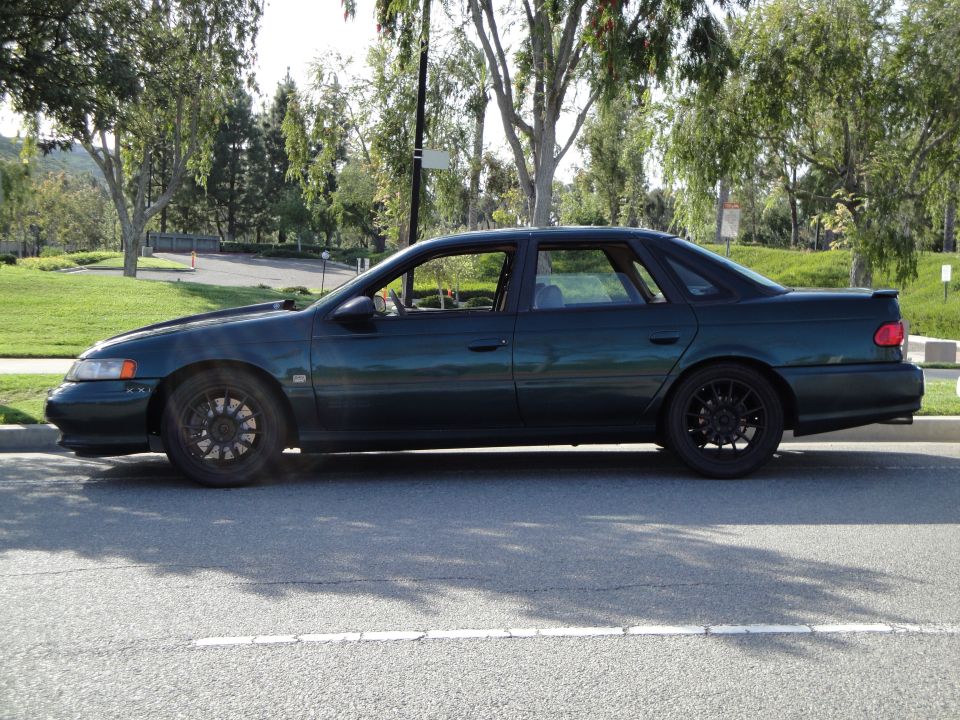 Dark green 1995 Ford Taurus sedan, side view, with black aftermarket wheels and a subtle rear spoiler