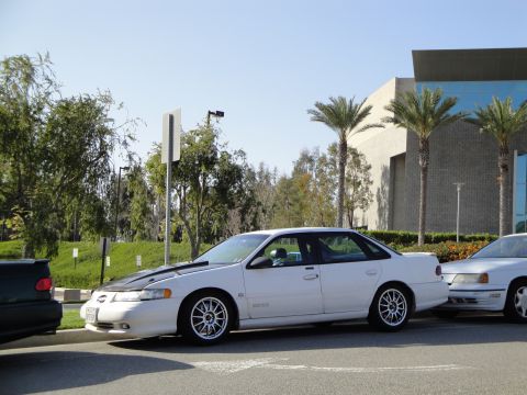 1995 Ford Taurus - Side view of a white 1995 Ford Taurus with a black hood and aftermarket wheels, parked next to another car.