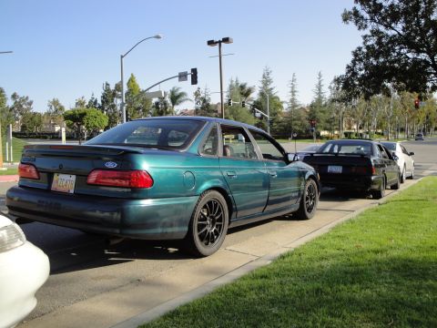 1995 Ford Taurus - A green 1995 Ford Taurus is parked alongside other vehicles on a sunny day.