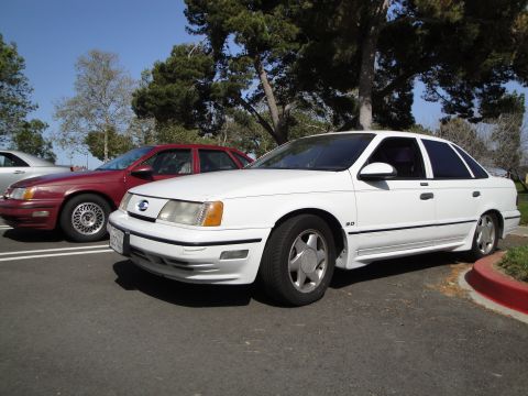 1995 Ford Taurus - White sedan parked next to a red sedan under trees.