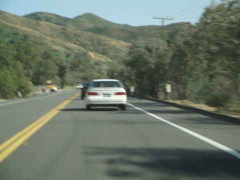 1995 Ford Taurus - A white sedan is seen from behind on a two-lane road, with mountainous terrain and trees lining the roadside.