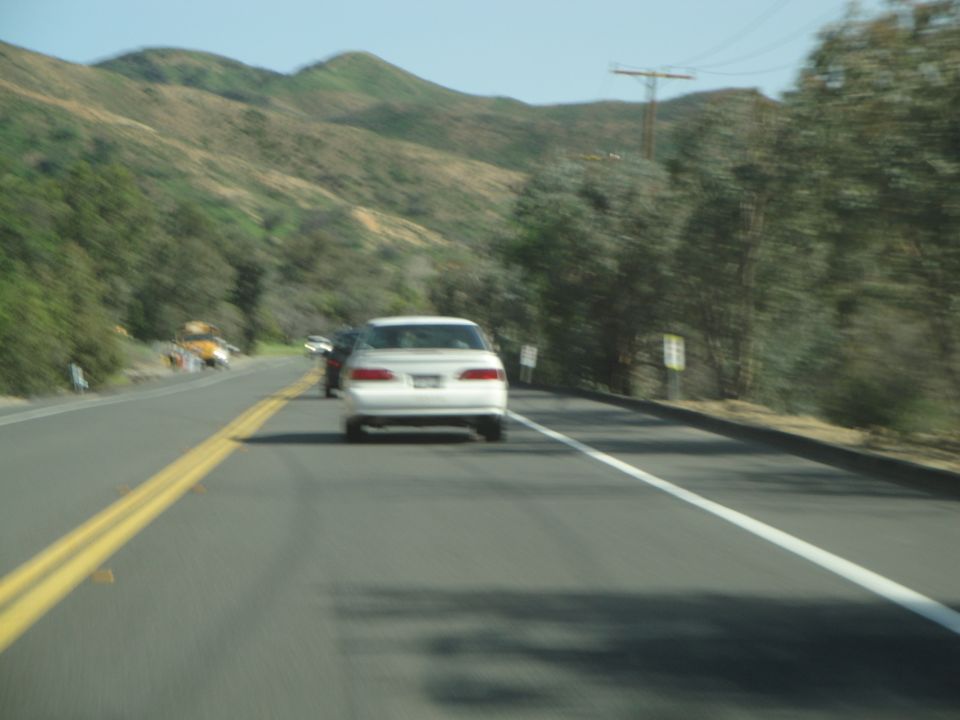 White sedan driving on a highway with hills and trees in the background. Another car is visible ahead