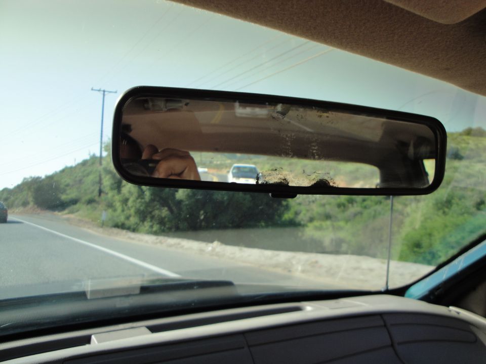 View from inside a car, looking through the windshield at the rearview mirror. Another car is visible in the distance on the road
