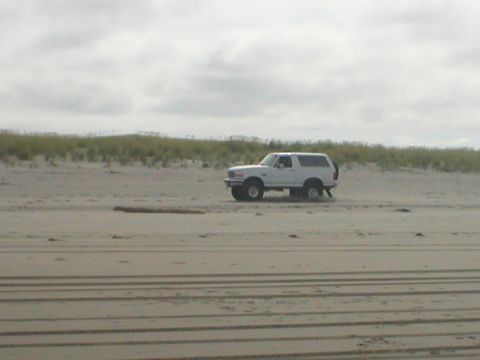 1978 Ford Bronco - White SUV with spare tire on rear door drives on a sandy beach under a cloudy sky.