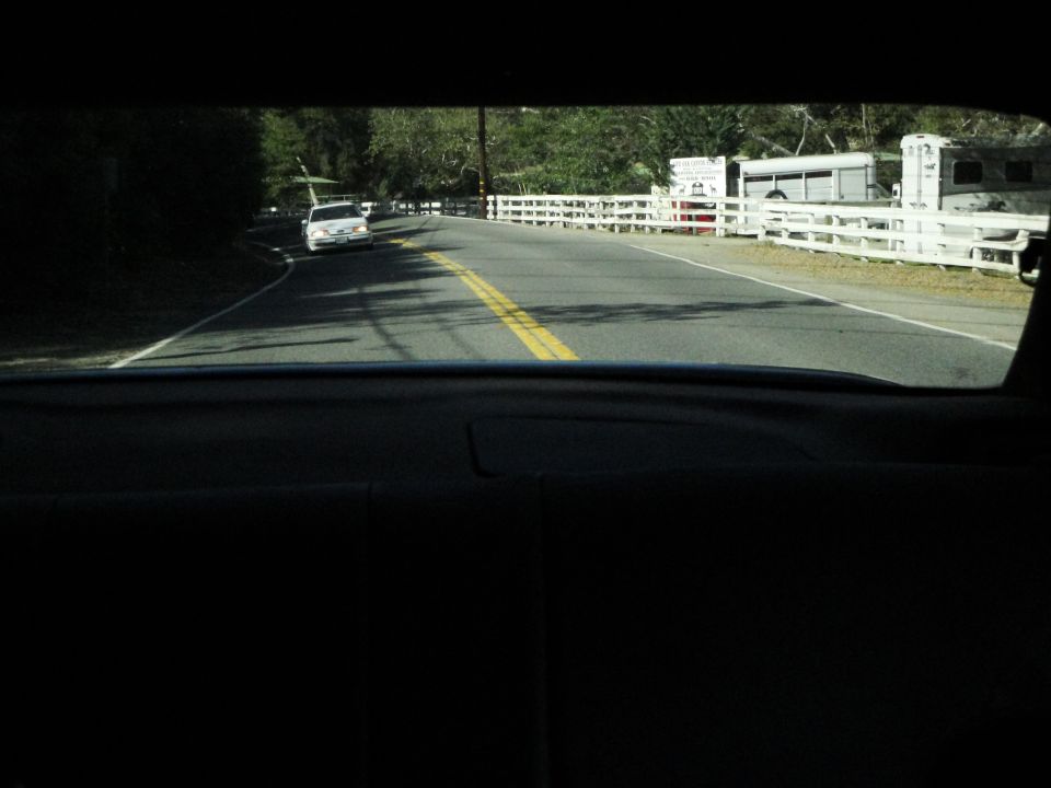 White sedan driving on a two-lane road with a white fence and horse trailers visible