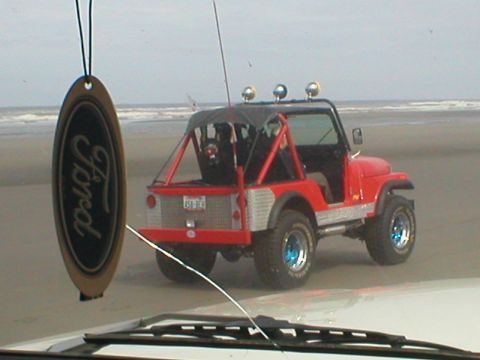 1978 Ford Bronco - A red off-road vehicle with a roll cage is parked on a sandy beach, with a Ford air freshener visible in the foreground.