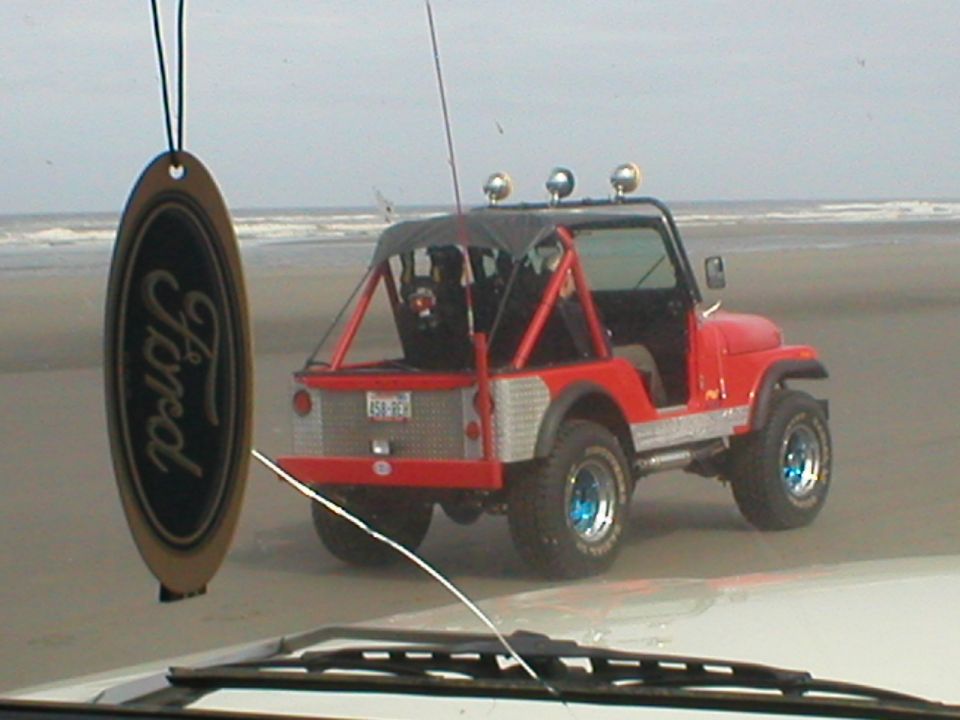 Red off-road vehicle with roll cage on beach; Ford air freshener hangs in foreground