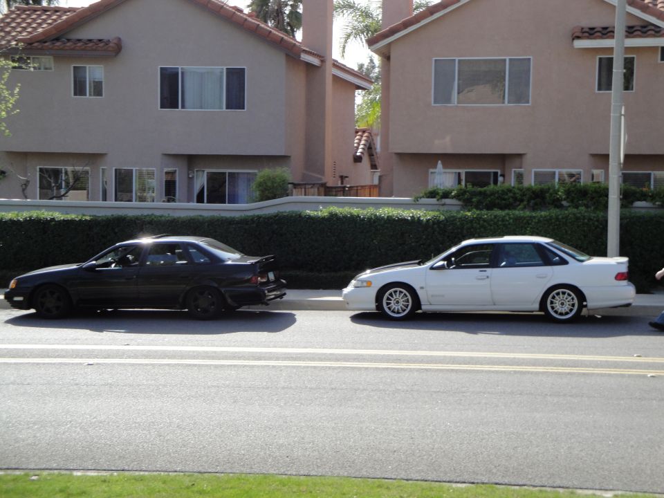 White sedan with black hood and aftermarket wheels parked next to a black sedan with a spoiler