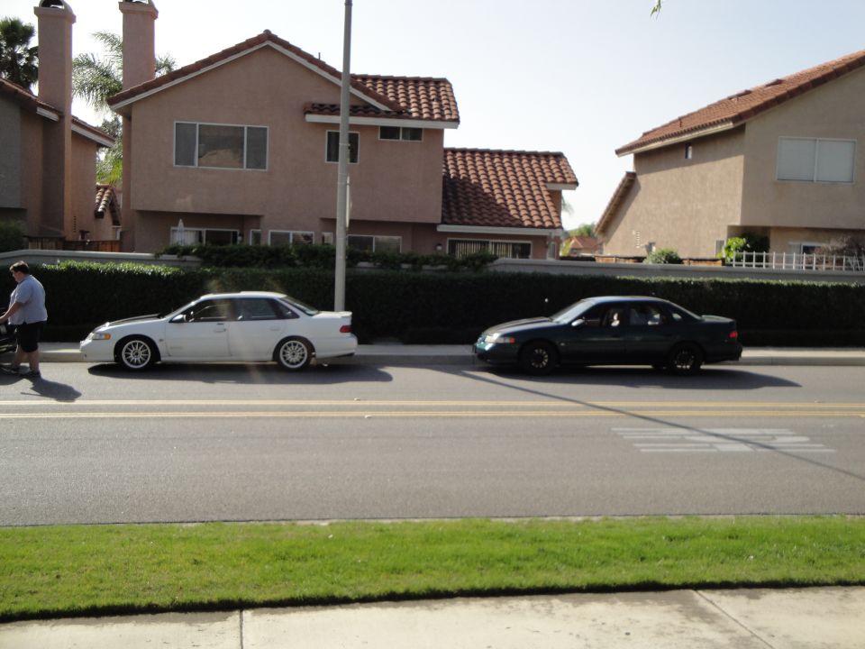 White sedan and dark sedan parked side-by-side on a street in front of houses