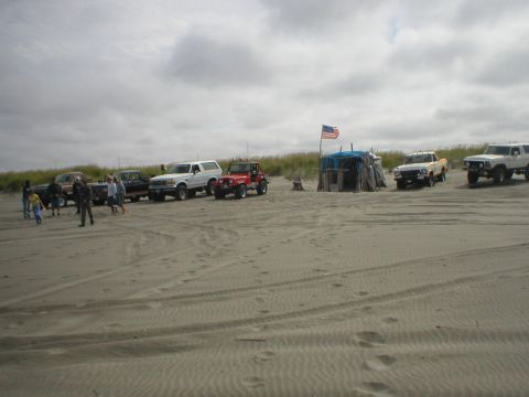 1978 Ford Bronco - A group of off-road vehicles are gathered on a sandy beach under a cloudy sky.