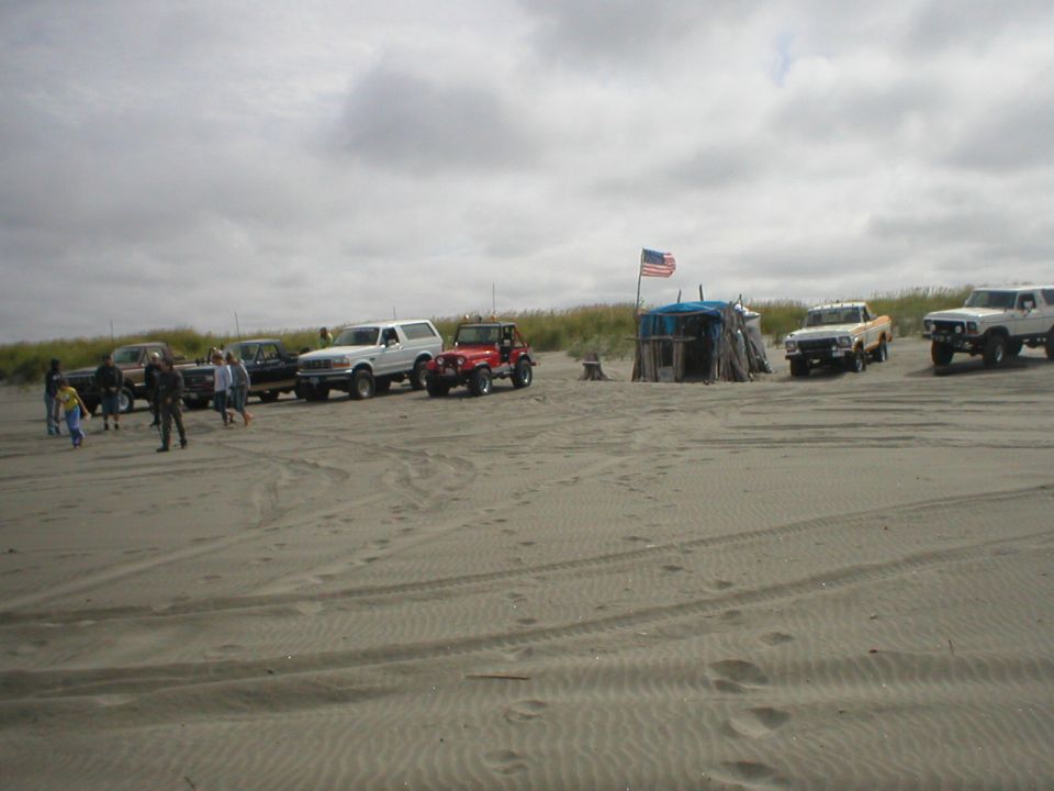 Several SUVs and trucks parked on a sandy beach with a small shack and American flag