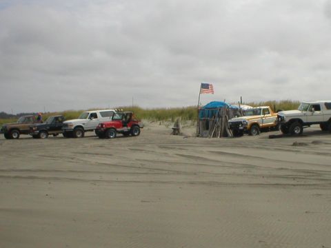 1978 Ford Bronco - Several SUVs and trucks are parked on a sandy beach near a rustic shelter flying an American flag.