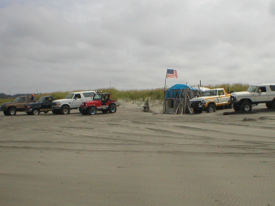1978 Ford Bronco and other vehicles on a sandy beach with a makeshift shelter and American flag