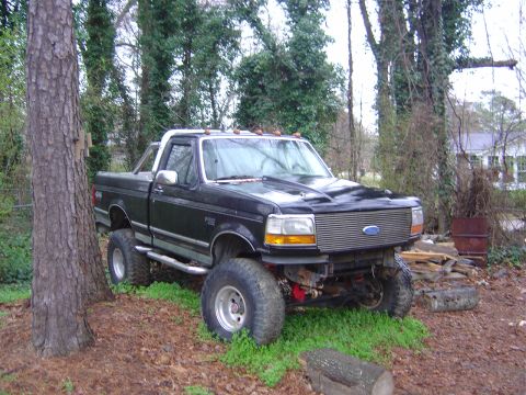 1986 Ford Bronco - A lifted black pickup truck sits in a wooded area with a chrome grille and large tires.