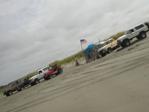 1978 Ford Bronco - A line of off-road vehicles, including SUVs and trucks, are parked on a sandy dune under a cloudy sky.