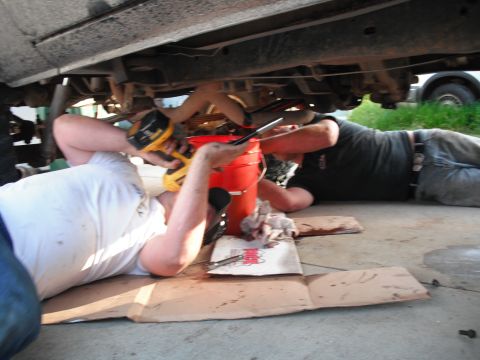 1991 Ford Bronco - Two individuals are seen working on the undercarriage of a vehicle, with tools and a bucket nearby.