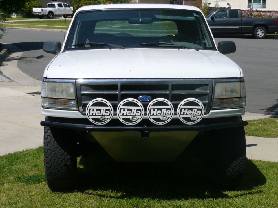 White Ford Bronco front end with four Hella lights on the grille and a custom bumper. A white pickup truck is parked in the background