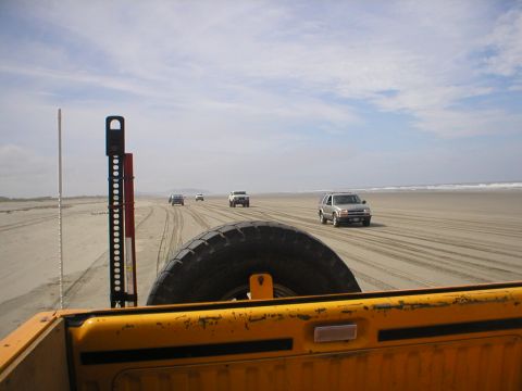 1978 Ford Bronco - Several vehicles drive along a sandy beach under a cloudy sky, viewed from the back of a yellow pickup truck.